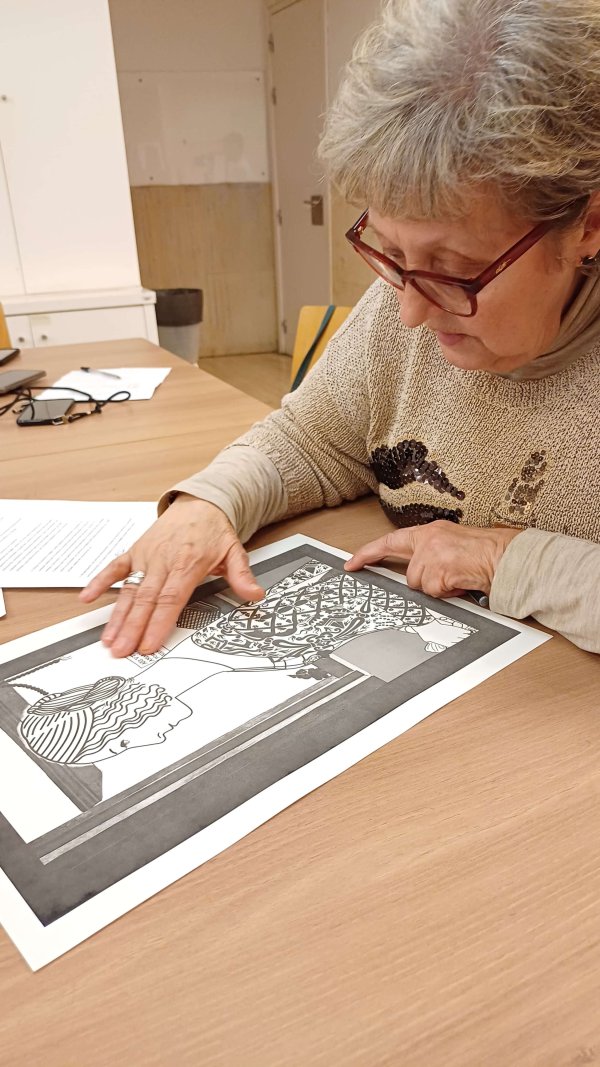 Mujer mayor con gafas observando y tocando una ilustración en blanco y negro sobre una mesa, mientras analiza sus detalles en una sala de reunión.